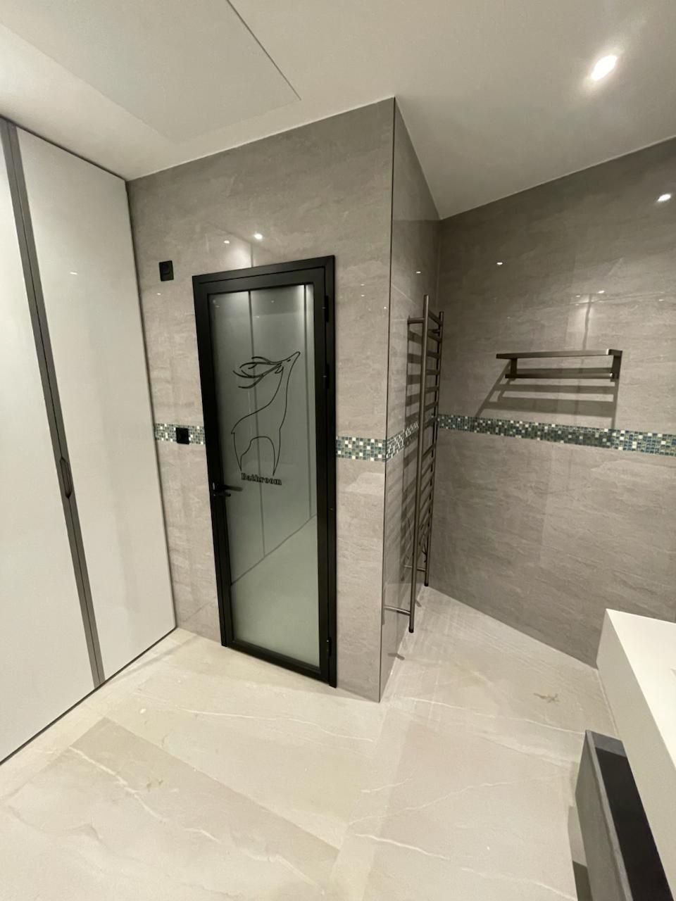 Grey porcelain-tiled bathroom with a black-framed etched glass door, heated towel rail and mosaic tile band