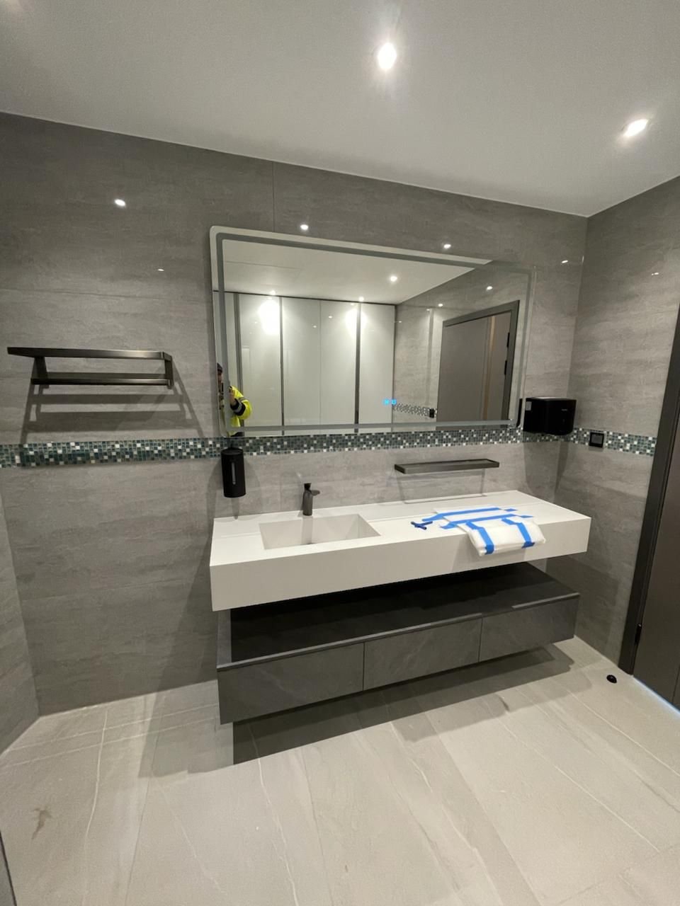 Floating white vanity with dark drawers below a wide mirror in a grey tiled bathroom