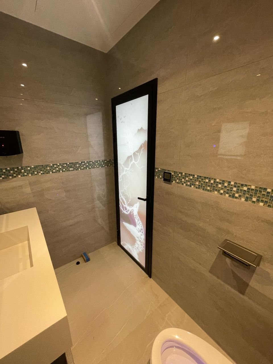 Black-framed glass door with a printed design in a grey tiled bathroom with a mosaic tile band