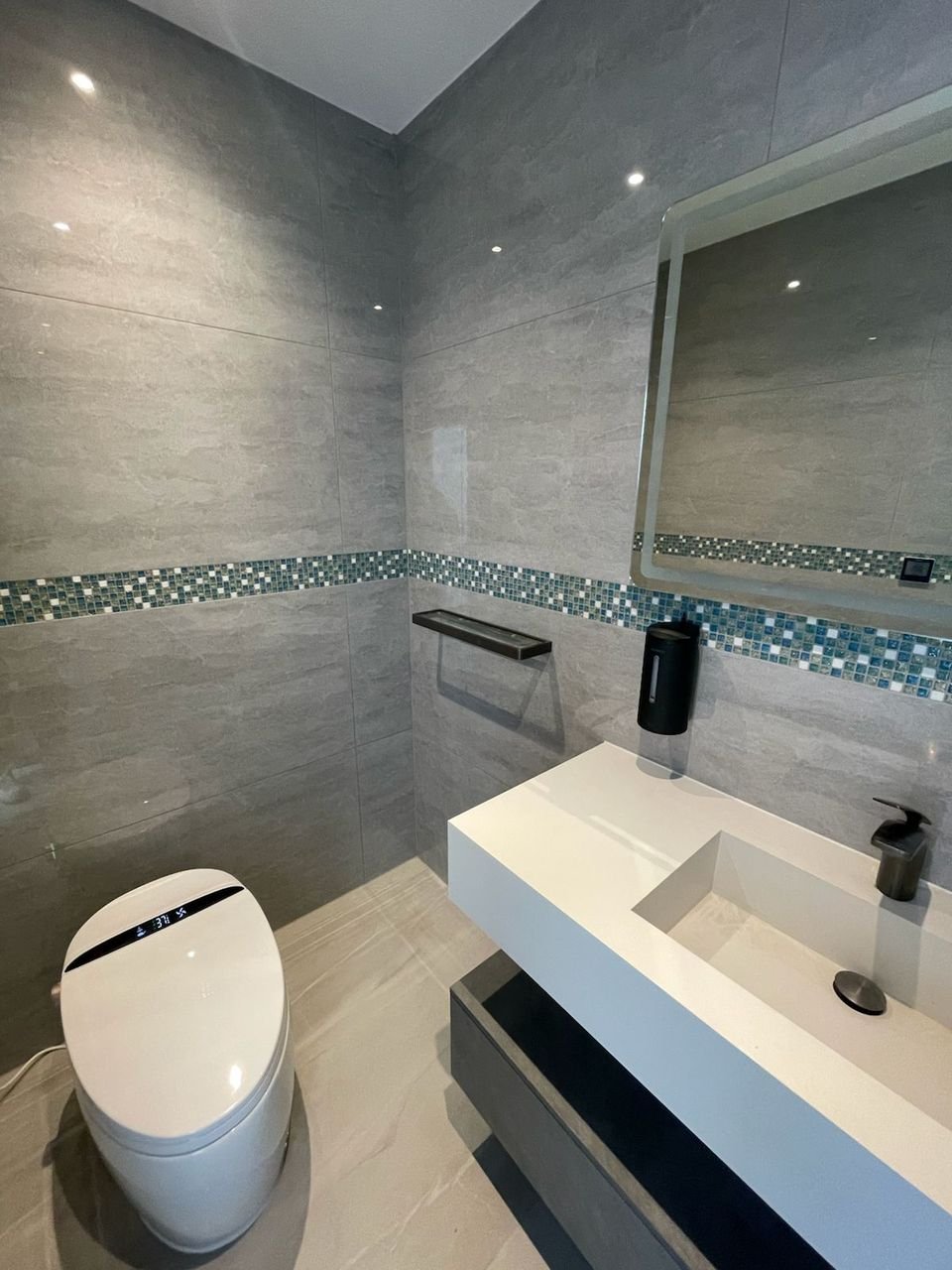 Smart WC beside a white basin, with a glass mosaic tile band and backlit mirror