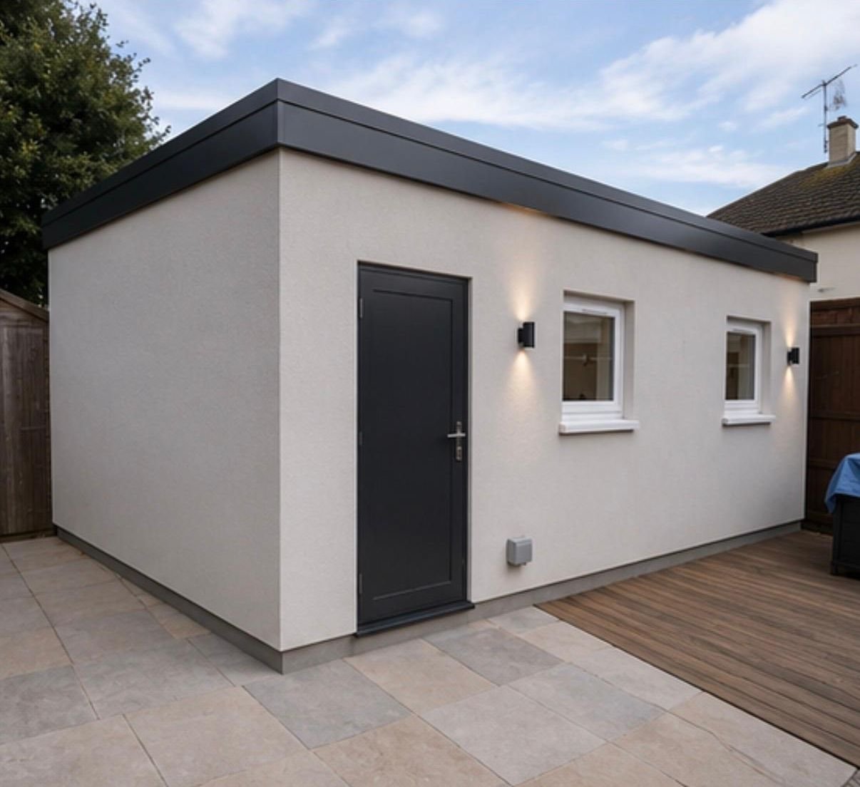 Rendered single-storey outbuilding with a flat roof, dark door and two windows beside a patio and deck