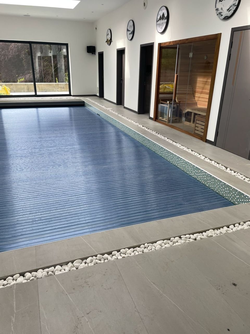 Indoor pool with a mosaic tile edge and slatted cover, with a glazed sauna and full-height windows beyond
