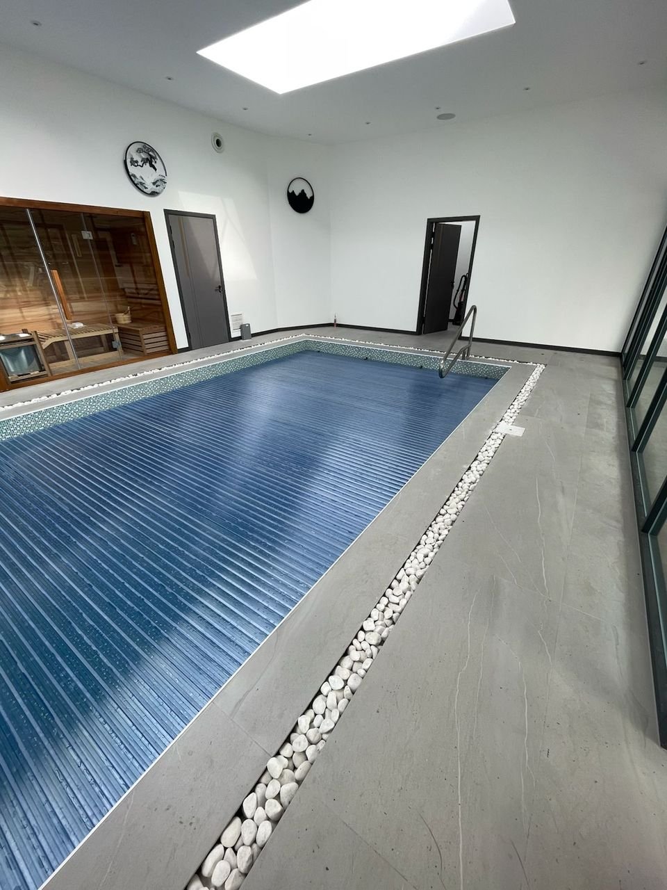 Indoor swimming pool with a slatted cover, porcelain surround edged with white pebbles, a rooflight and a glazed sauna
