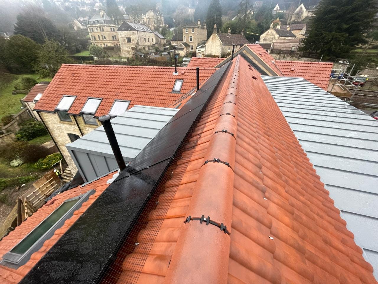 New clay tile ridge and aluminium roofing viewed from the roof, with stone houses and trees beyond