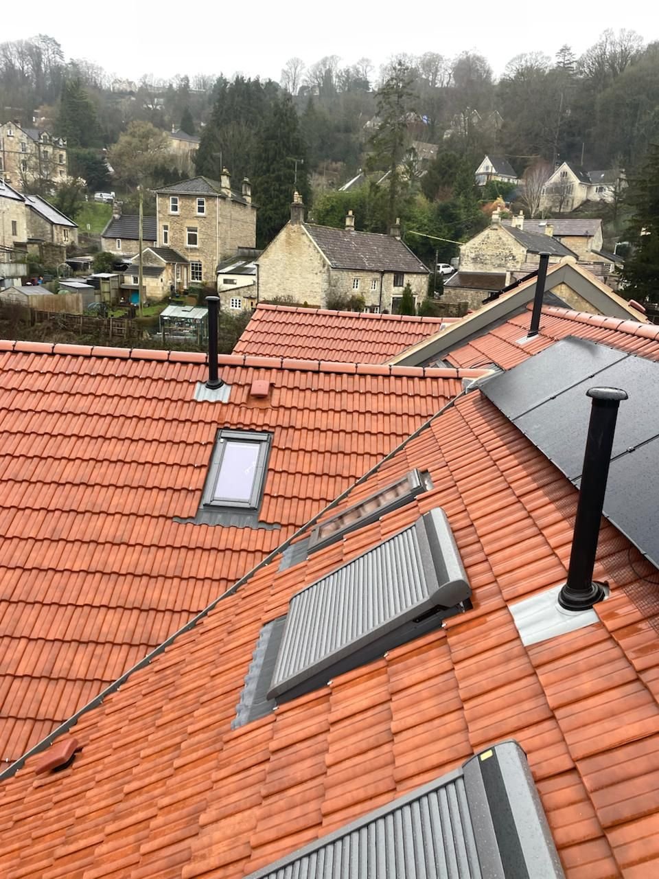 Clay tile roof with rooflights, flues and solar panels, looking out over stone houses on a wooded hillside