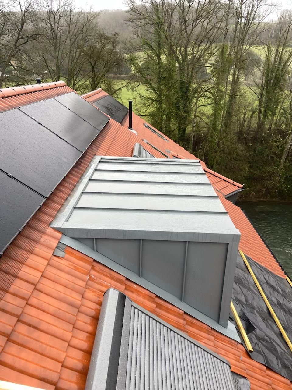 Aluminium-clad dormer with a standing seam roof set into a clay tile roof beside solar panels, with trees beyond